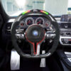 Front view of a BMW F chassis, M sport steering wheel in carbon fiber, hollow design paddle shifters, a mix of red and M color accents, black perforated leather and led shift lights